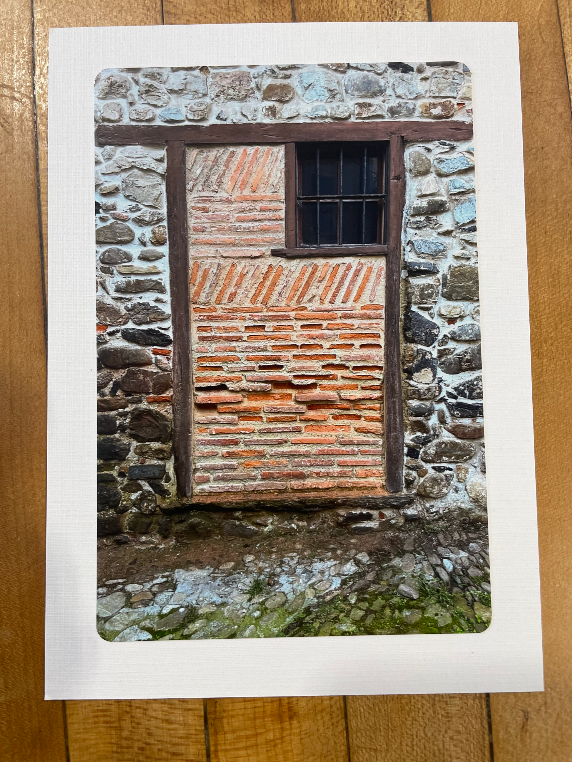 A square art print with a white border showing a textured painting of a rustic stone wall and brick window, displayed on a wooden surface.
