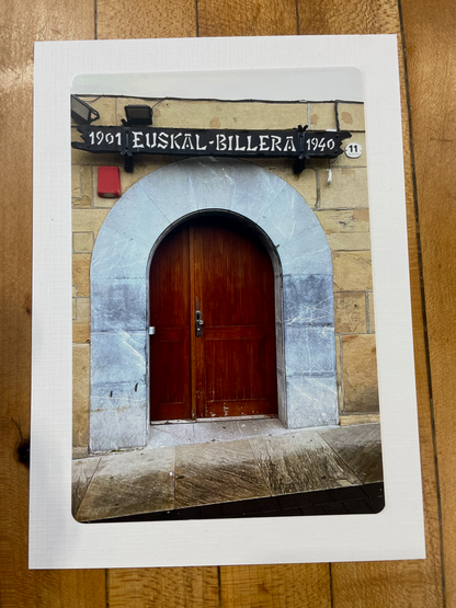 A printed postcard or photo print showing an arched wooden door set in a stone wall, photographed from above on a wooden surface. The print has a white border and a small black sign reading '1901 EUSKAL-BILLERA 1940' is visible on the printed image.
