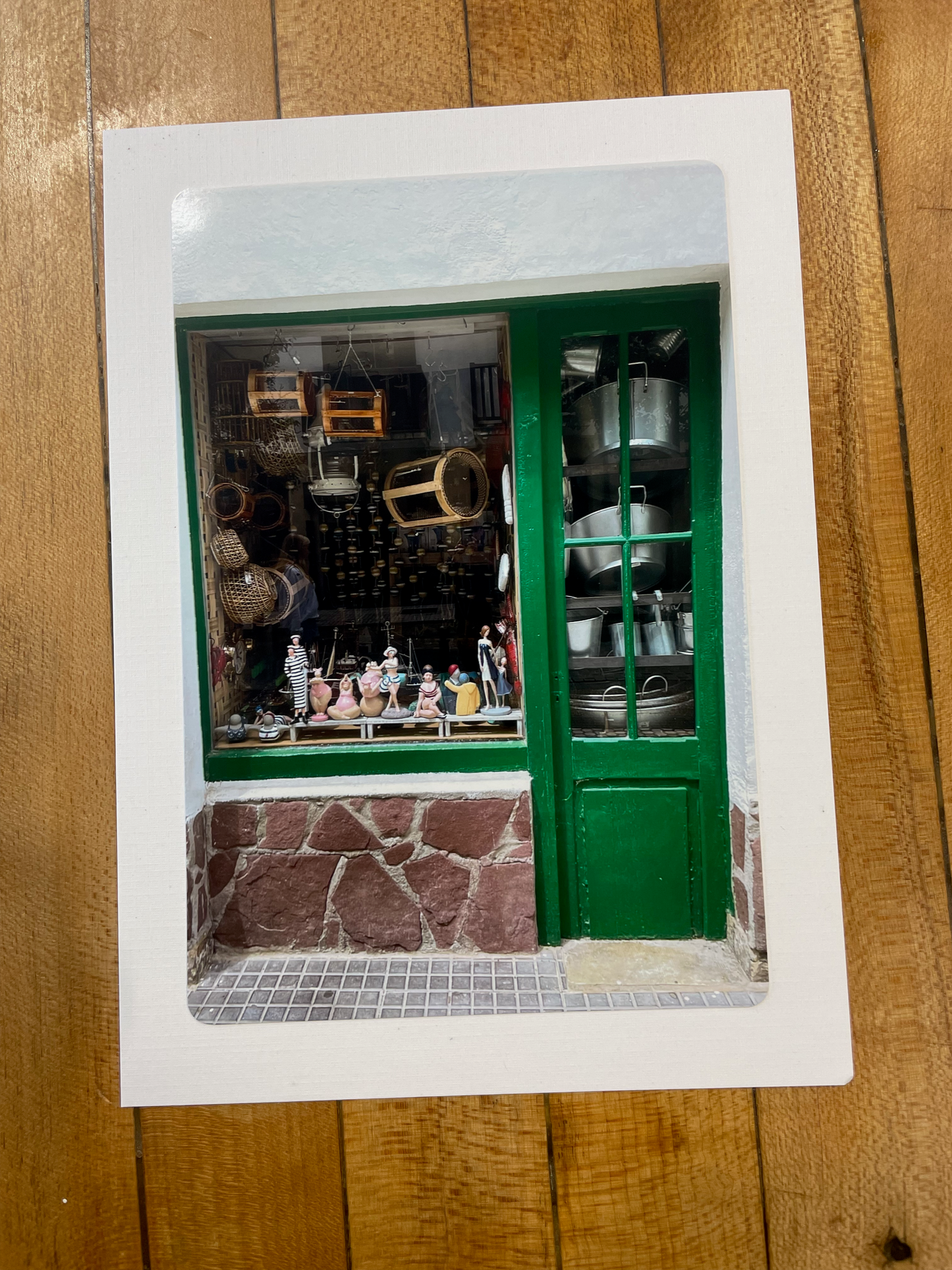 A square white art print or card featuring a photograph of a green storefront window with shelves of bottles and jars, placed on a wooden surface.