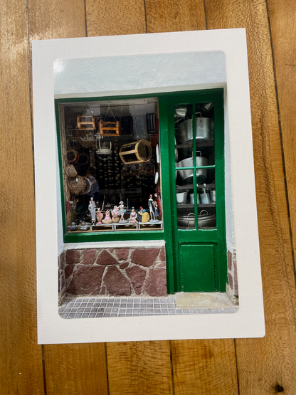 A square white art print or card featuring a photograph of a green storefront window with shelves of bottles and jars, placed on a wooden surface.