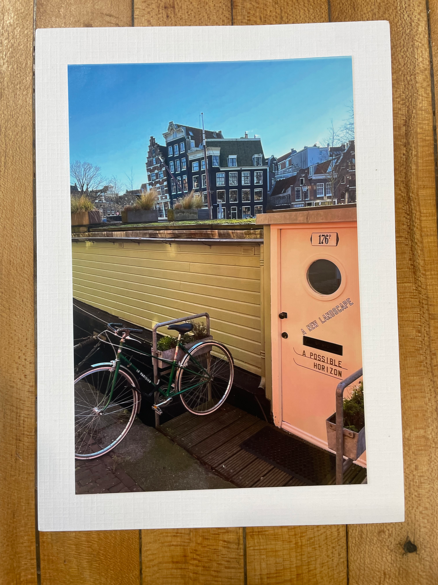 A square printed photo or postcard with a white border, placed on a wooden surface. The printed scene shows a canal-side street with bicycles, a pink mailbox, and blue-gray buildings under a bright blue sky.