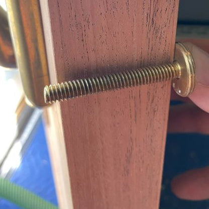 A close-up of a brass-colored threaded screw with a round flat head, shown fastened against a reddish-brown wooden surface, with a finger holding the side.
